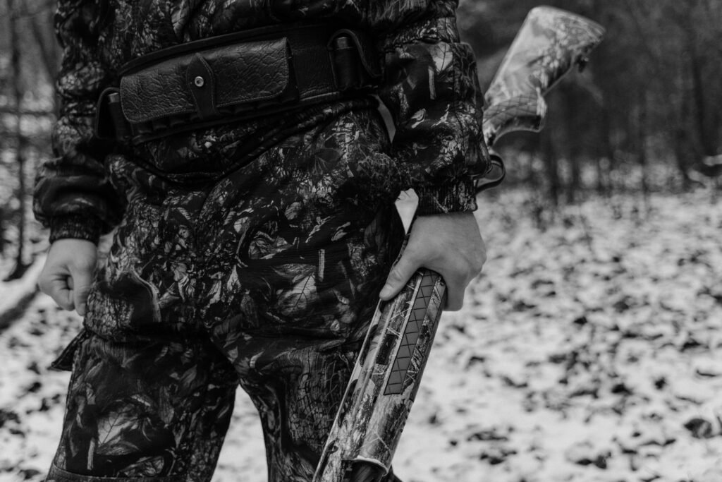 Monochrome image of a hunter in camouflage gear holding a shotgun in a snowy forest.