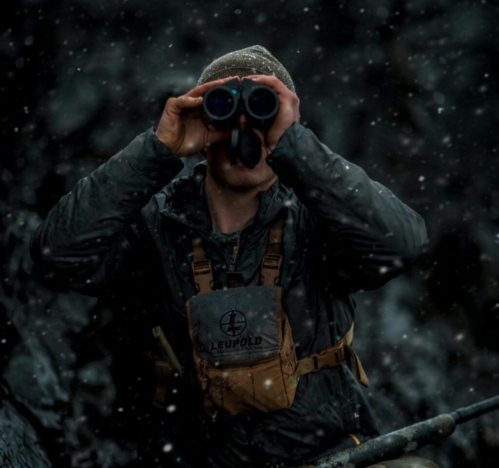 Hunter using binoculars in a snowy winter scene, rifle in hand.